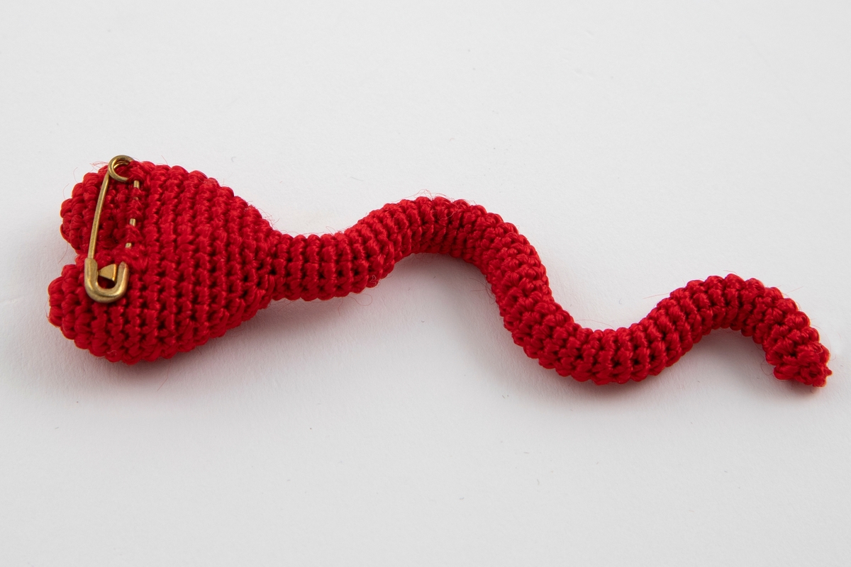 Heklet brosje i rødt garn med en bred, hjerteformet øvre del og en smal, slyngete nedre del som ligner en hale. Sikkerthetsnål på baksiden til å feste smykket.