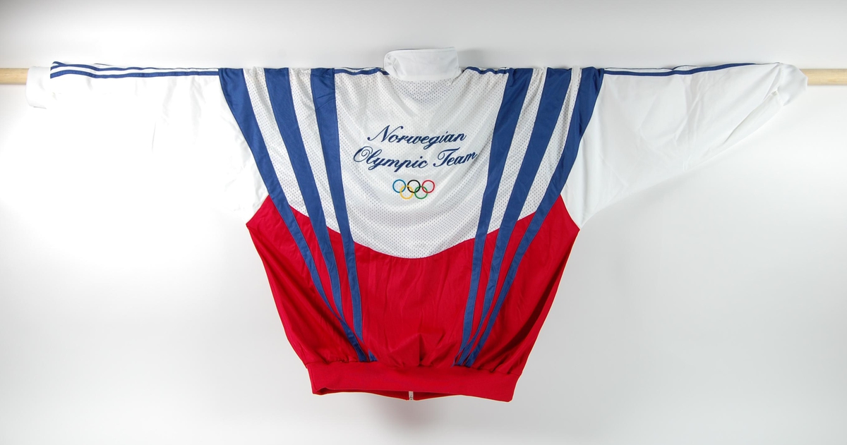 Overtrekksdress med hovedfargene rød, hvit og blå. Buksa er kun rød og hvit. Pålimt merke med logo for Olympiatoppen på jakka. OL-ringene er pålimt på ryggen.