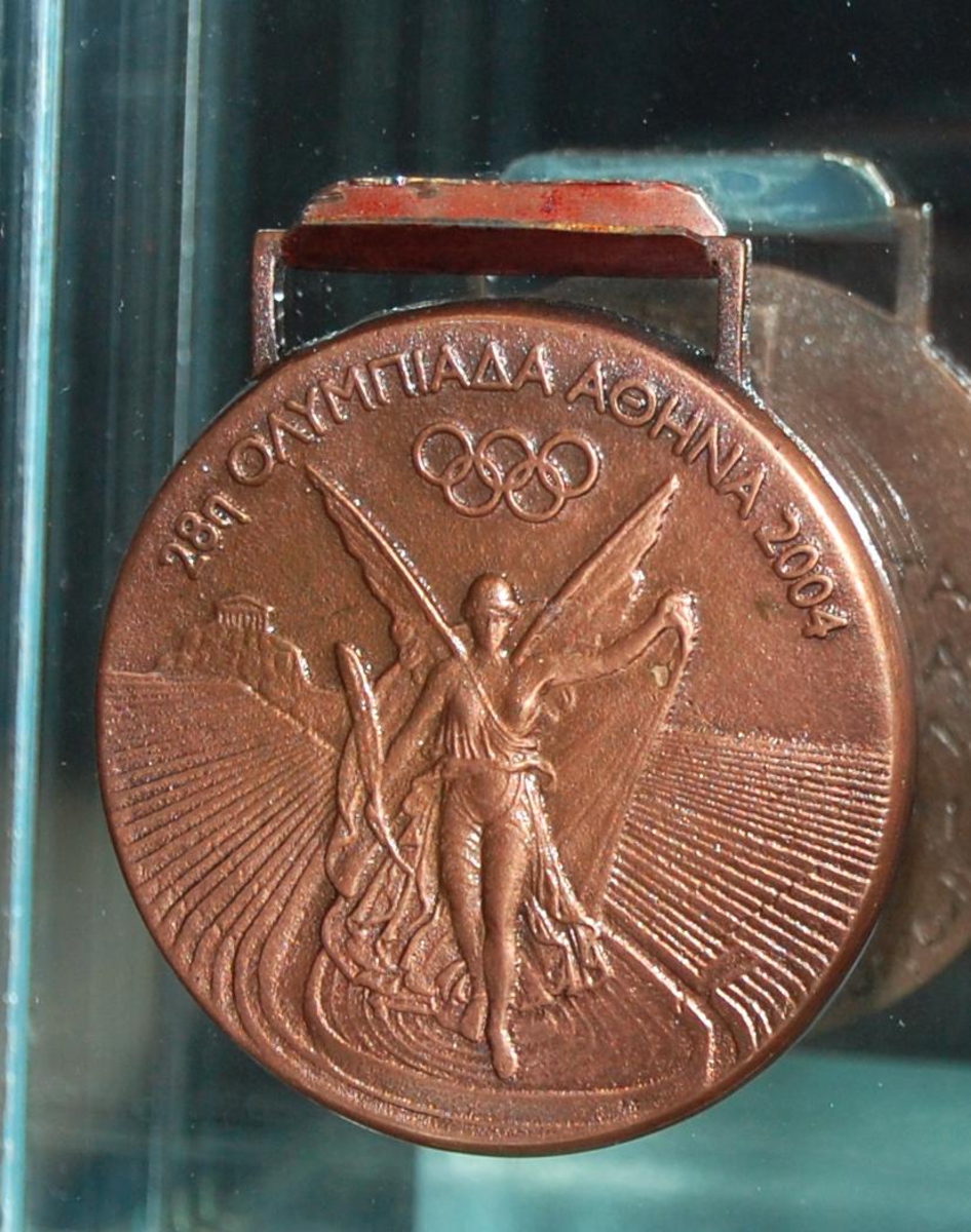 Gullmedalje med motiv av den greske seiersgudinnen Nike som flyr over en stadion. Medaljen har gresk innskrift.
