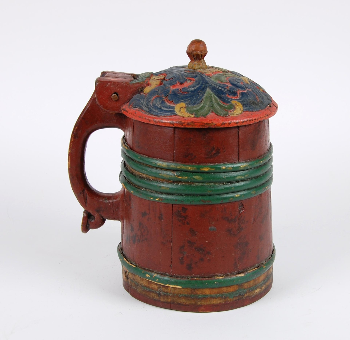 Kanne av furustaver. Lokk med akantusskurd malt i grønne, gule og blåe farger. Kuleformet lokkgrep. Korpus med fire grønnmalte gjorder oppe og en nede (opprinnelig tre). Hanken er utskåret av en av stavene, ender nederst i en plate med bladmotiver.