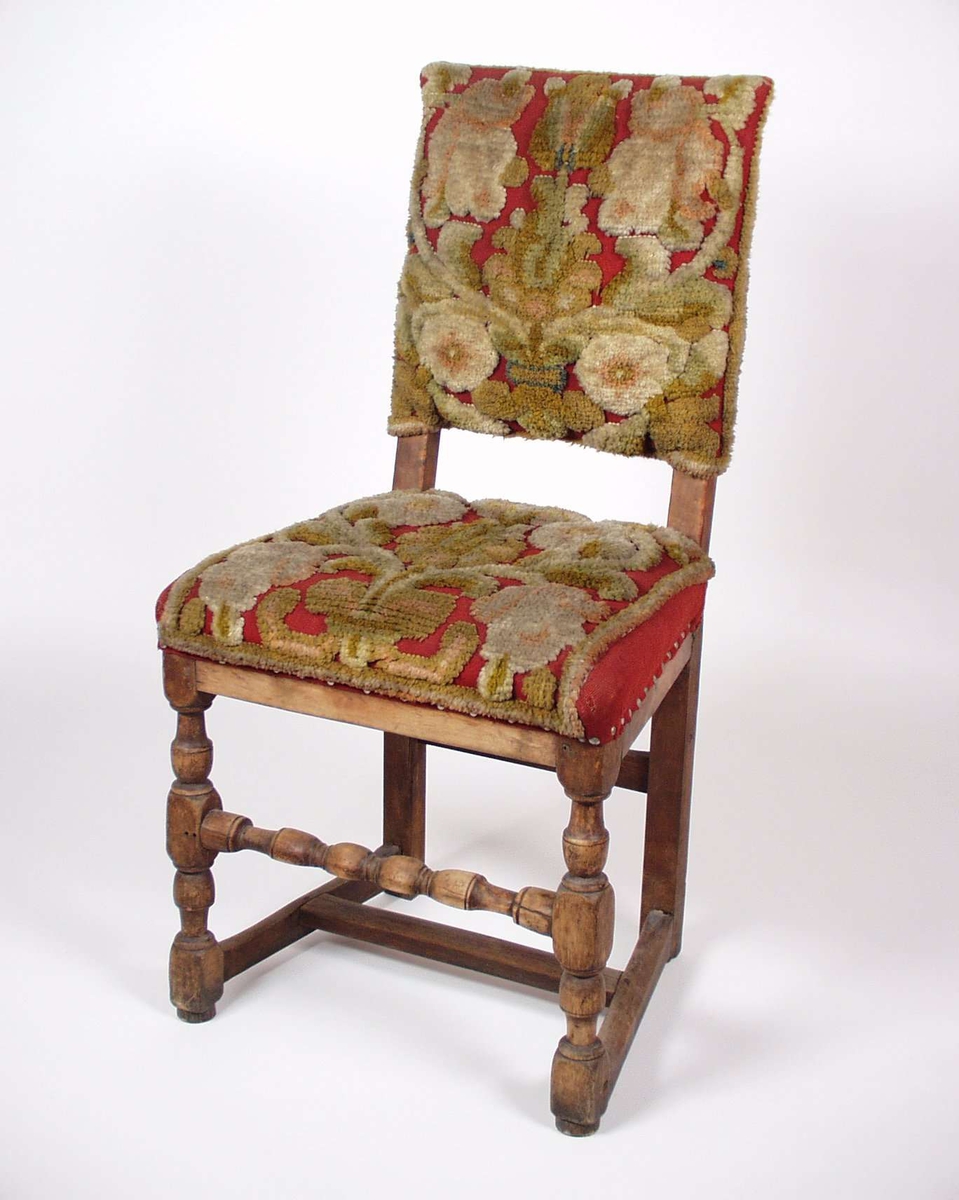 Stolen er av tre med mønstret rødt tekstiltrekk. Midtspross og forbein er dreid.
