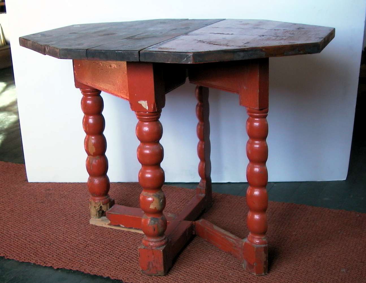 Åttekantet klaffebord med fire ben og benforsterkning. Bordet er rødmalt. Bordplaten er stygg. Treverk er slått av på foten. Malingen er avslitt.