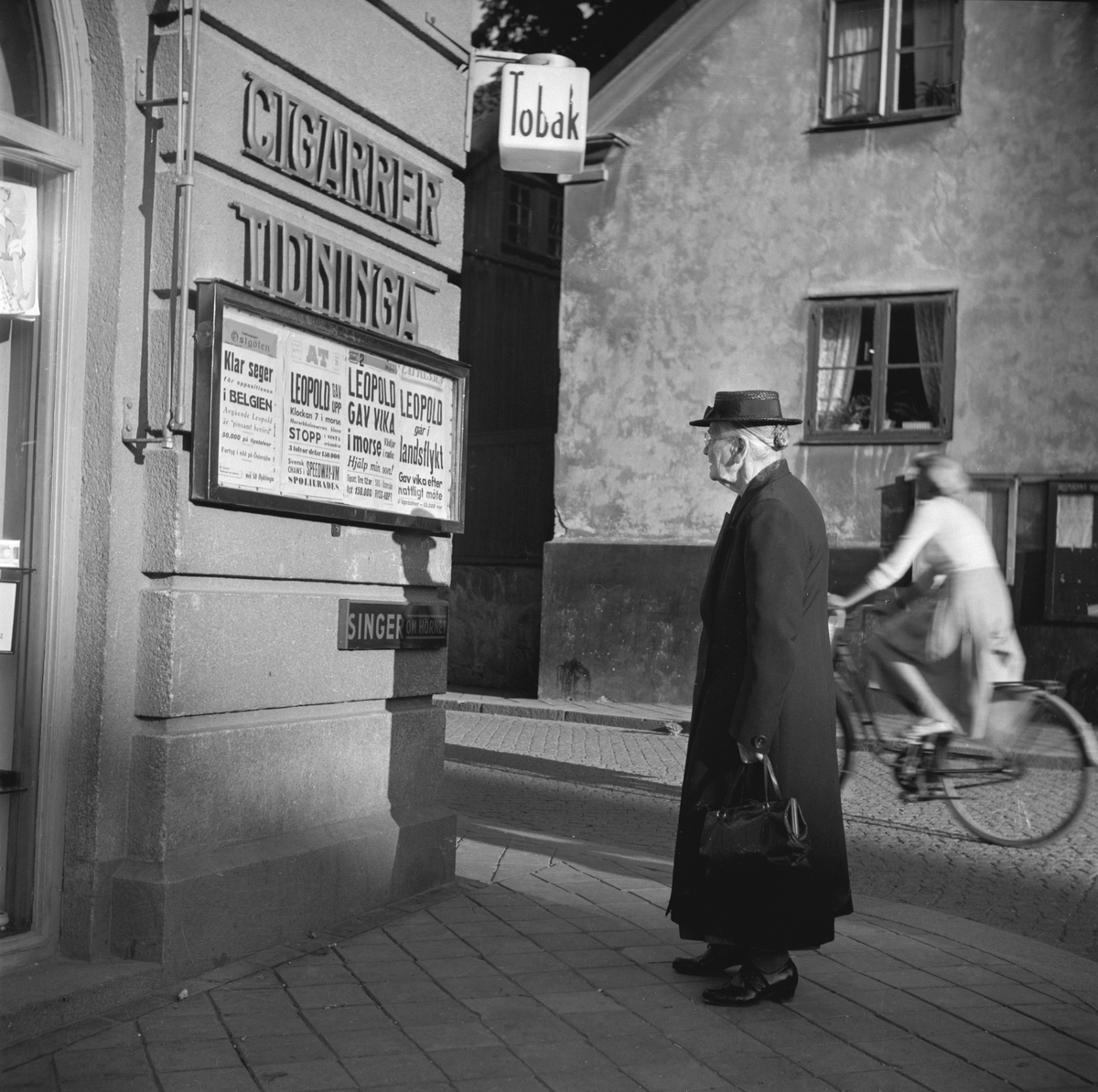 Tobakscentralen i hörnet av Stora Torget och Tanneforsgatan i Linköping. En äldre dam läser kvällstidningarnas löpsedlar. Likt många andra svenskar följde hon sannolikt med intresse utvecklingen av efterkrigsårens konstitutionella oro i Belgien. Kung Leopold III var ju änkling efter den mycket populära svenska prinsessan Astrid, som så tragiskt omkommit i en bilolycka. Denna septemberdag 1951 berättade löpsedlarna om den belgiska krisens upplösning. Kung Leopold avsade sig kronan till förmån för sin äldste son, kronprins Baudouin. Upplysningsvis drevs Tobakscentralen fram till 1957 av Bertha Palmqvist. Därefter innehades den av Lennart Danell.