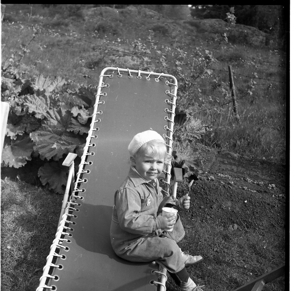 En liten flicka sitter på en solstol med en sked och en kopp i händerna. Hon har en liten mössa på huvudet.