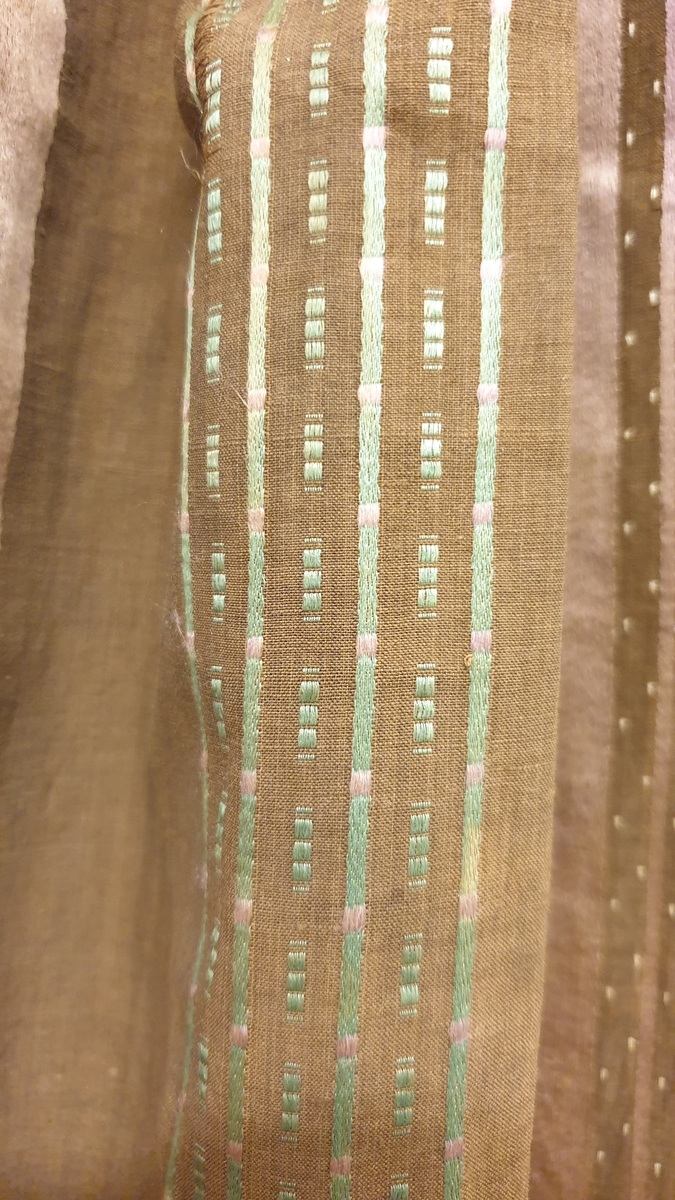 Förkläde i brun bomull med extra mönstervarp i lila (nu rosa), grönt (blekt) och ofärgat silke som flotterar på fram-och baksida. Mönster i form av prickar och fyrkanter samt ränder i bredar ränder i varpsatin?/kypert. Bredd i midjan 18 cm. Handsytt.

Enligt uppgift på registerkort saknas linning och knytband, men förklädet har ett svart ripsband fastsytt som midje- och knytband.