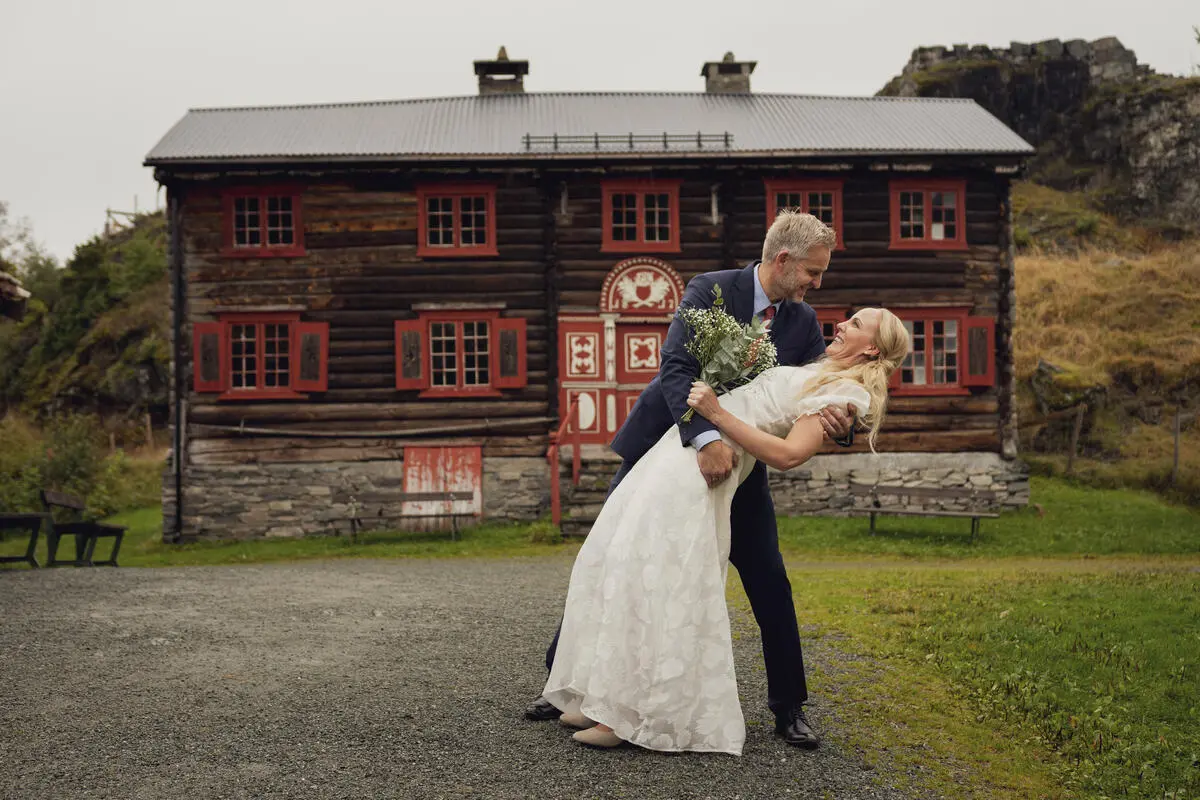 Et brudepar holder rundt hverandre foran gammelt hus. Foto. 