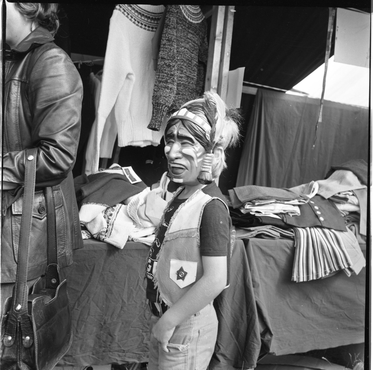 En pojke med comboyväst och indianmask står vid ett marknadsbord med kläder.