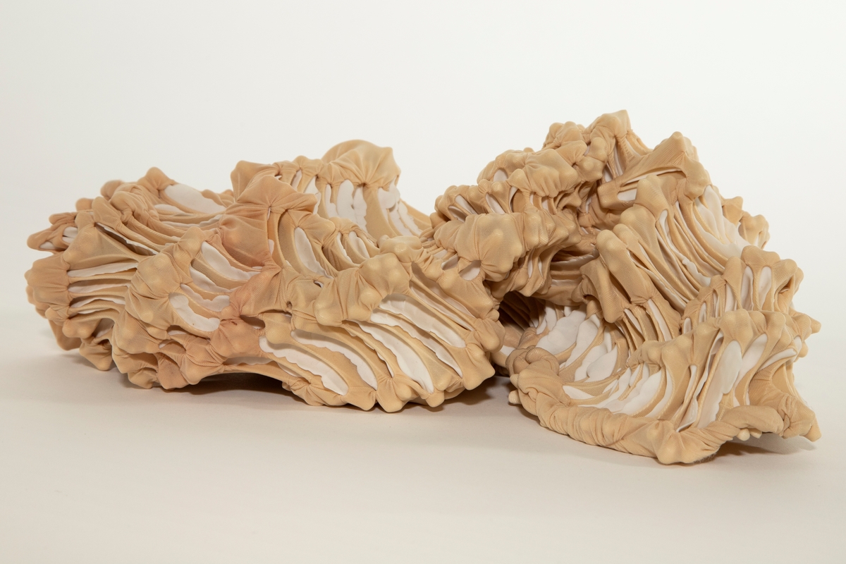 Objekt/ skulptur med organisk form. Beige nylon er knyttet og sydd rundt porselensskiver og foldet. Formen kan minne om et kadaver.