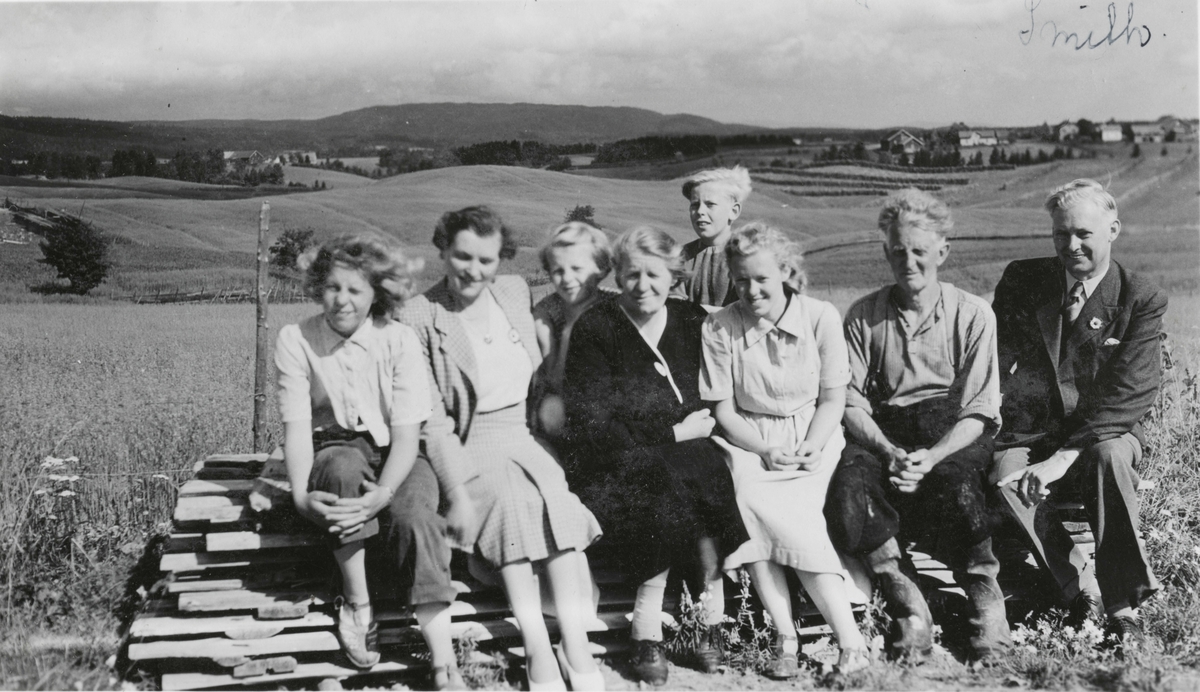 En gruppe mennesker sitter på plankestabel. Utsikt over jorder i bakgrunnen. Gjerdrum.