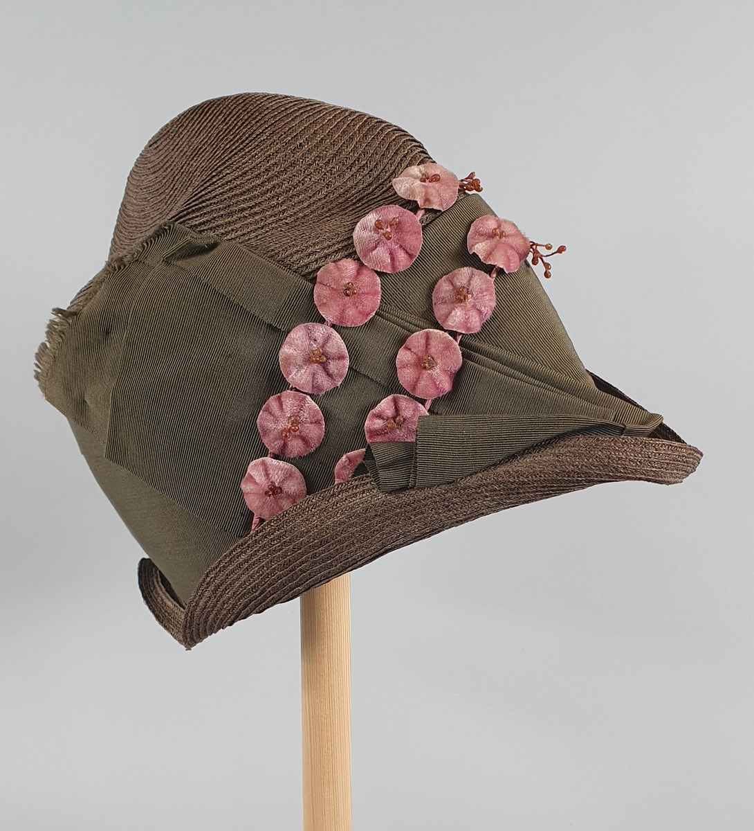 Brun damehatt av strå, med høy pull og smal brem som er brettet opp foran. Bredt, mosegrønt hattebånd med rosa blomster rundt pullen. Hatten er delvis fôret på innsiden.