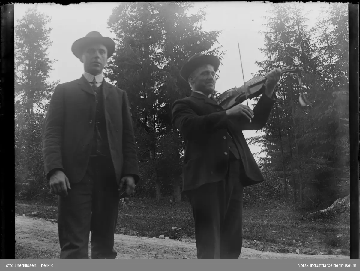 To menn stående på vei med skog i bakgrunnen. Begge kledd i dress og hatt. En av mennene spiller på fele.