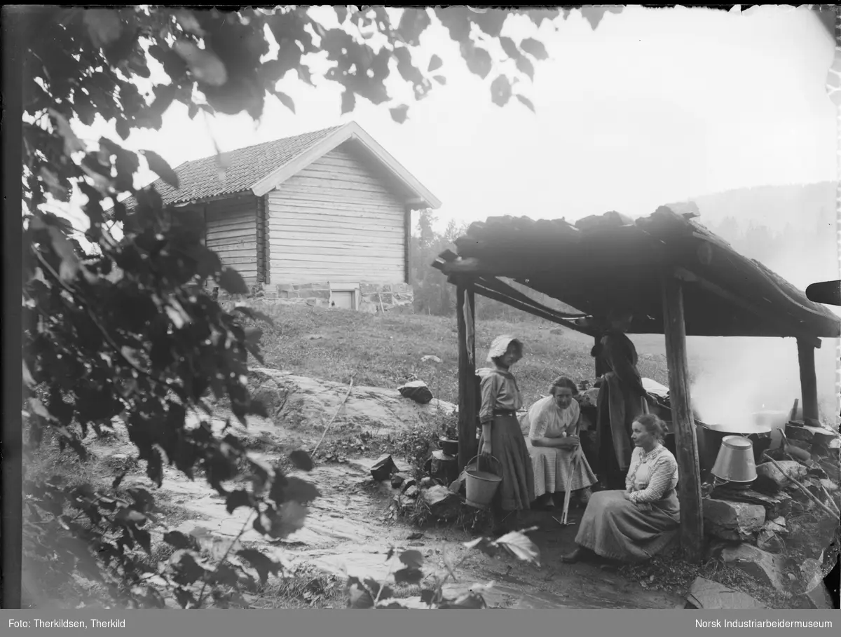 Fire kvinner sittende under overbygget vaske- eller melkeplass på Tovestul. Den ene kvinnen rører i bryggerpanna.
