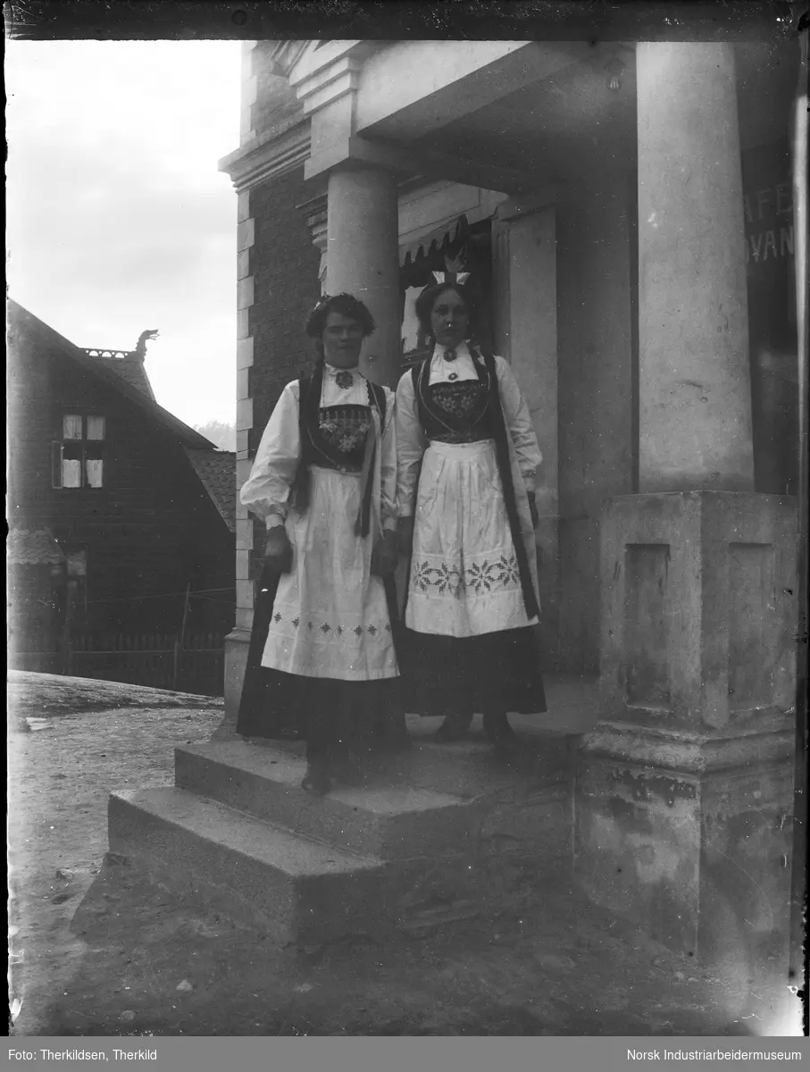 To kvinner i bunad stående på trappen inn til Trudvanggården. Bygningen er i mur, murt overbygg over trappen med søyler ned. "Håndgjerningsskolen" til venstre.