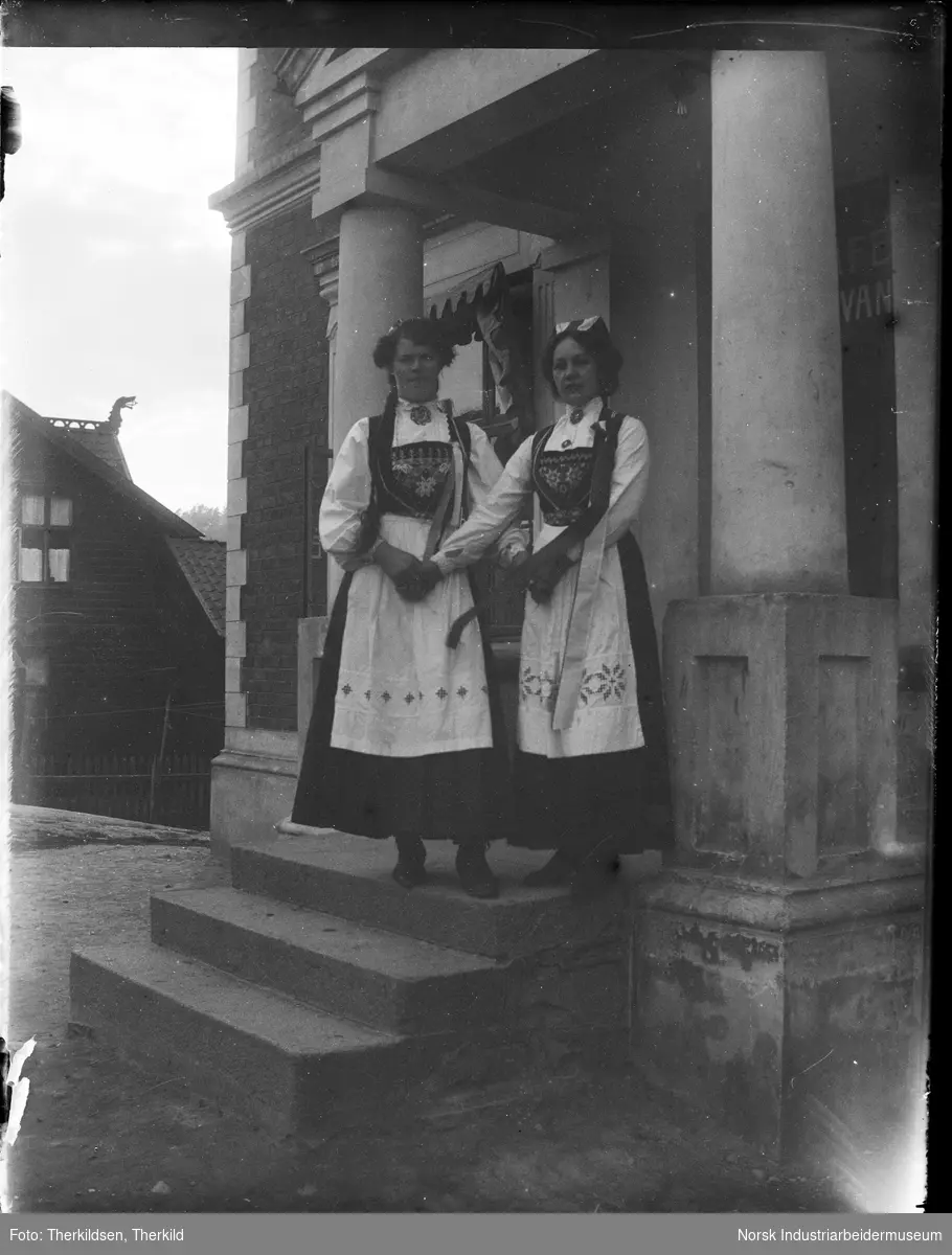 To kvinner i bunad stående på trappen inn til Trudvanggården. Bygningen er i mur, murt overbygg over trappen med søyler ned. Kvinnene holder hverandre i hendene. "Håndgjerningsskolen" til venstre.