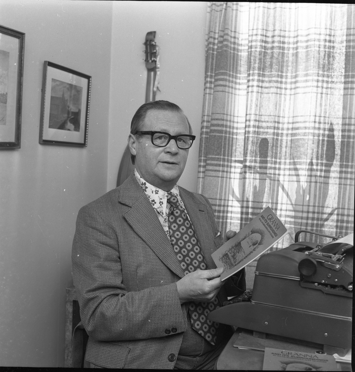 En man i smårutig kostym med prickig slips och blommig skjorta sitter vid ett bord med skrivmaskin och håller upp en skrift: "Gränna and its surroundings." På väggen bakom honom hänger troligen en luta.