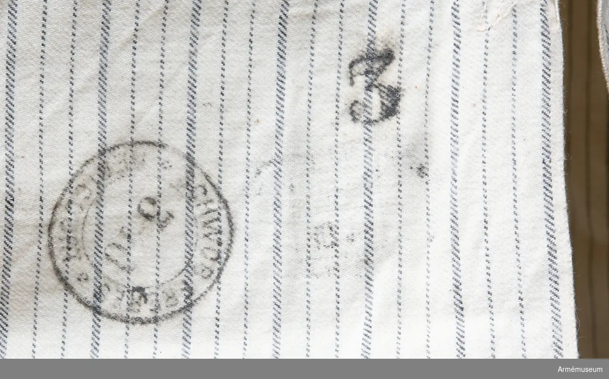 Skjorta av bomullstyg (vit med gråa ränder) med fast upprättstående krage och långa ärmar. På bröstets öppning två knappar, och på krage en; alla äro små, vita av ben. Likadana knappar, en å varje, på ärmuppslagens sprund. På skjortans framsida, en stämpel: "104de L" - 104 linjeinfanteriregementet. På skjortans baksida stämpel "30.11. 1910" - allt annat oläsligt.