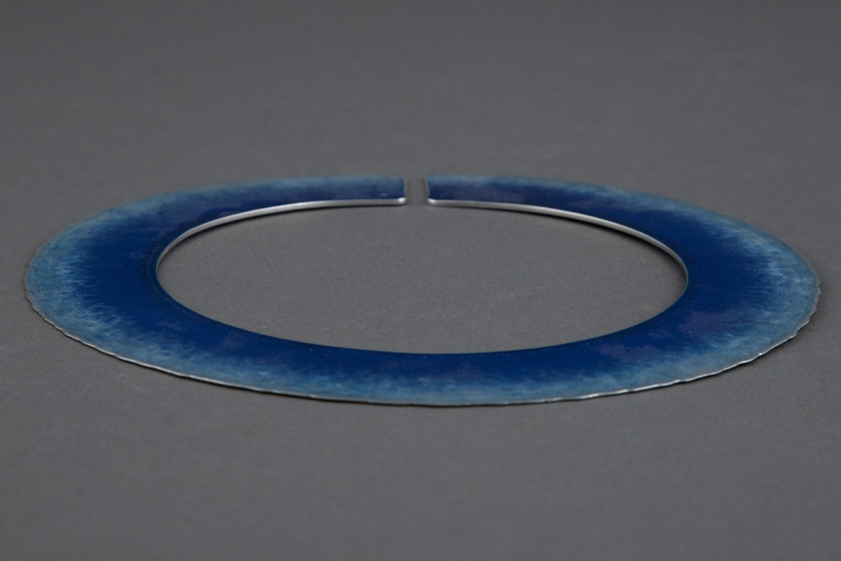 Halssmykke bestående av en cirka 3 cm bred aluminiumsplate i sirkelform med en liten åpning. Begge sider av platen har en dyp mellomblå farge innerst. Fargen blir gradvis lysere mot yttersiden, og den ender i en svakt, bølgeformet kant ytterst.