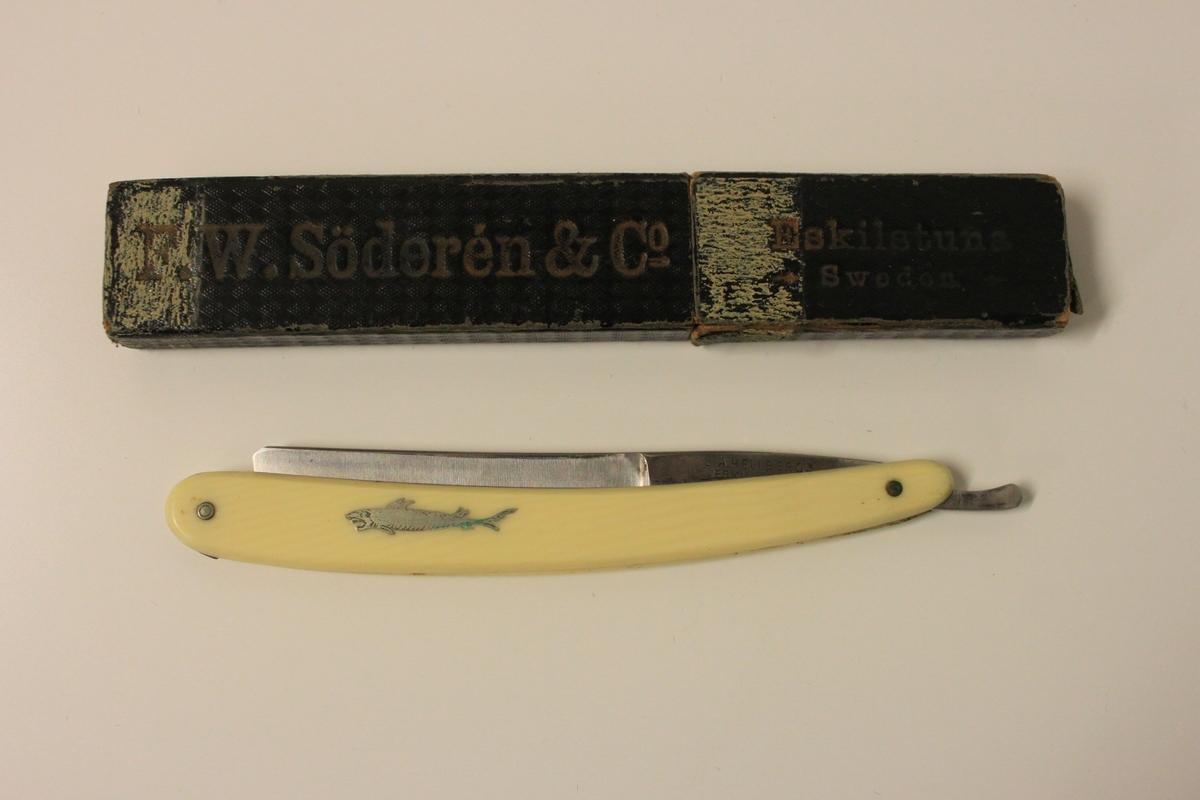 Rakkniv försedd med ett gulnat benskaft och en haj som motiv. Etui i svart med texten F.W. Söderén & co, Eskiltuna Sweden och grön insida.
