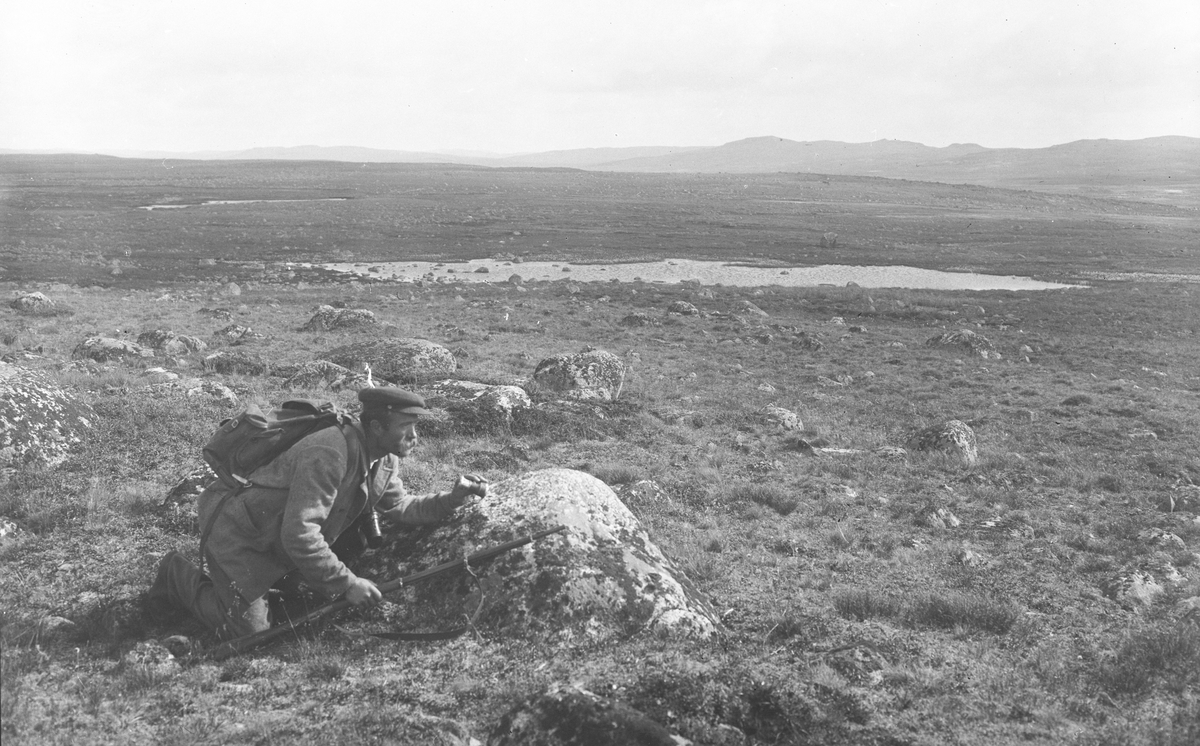 Reinjeger Sylfest Sæbo ligger i dekning bak en stein med kikkert og gevær for hånd, 1914.