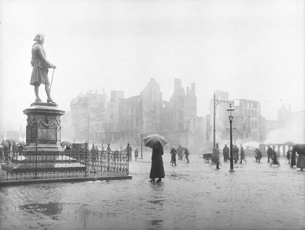 Ludvig Holberg-statuen og mennesker med paraplyer på Torgallmenningen i Bergen foran rykende brannruiner etter bybrannen 15. januar 1916.