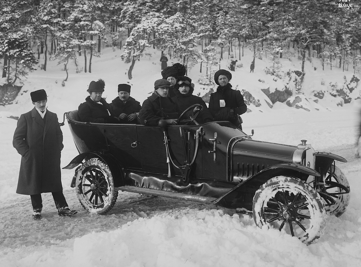 Tre kvinner og fire menn i åpen bil i vinterlandskap. Ifølge protokoll tilhører bilen en pølsemaker Christiansen fra Christiania. Fotografert 1912.