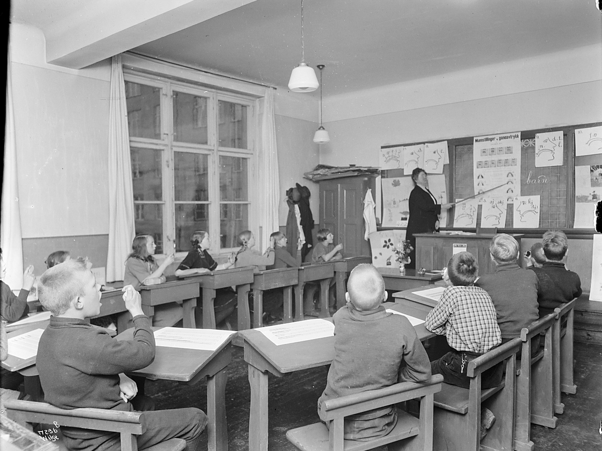 Elver sitter ved pultene sine og ser på lærerinne Jacobine Ulrica Rye står ved tavlen og underviser, Granhaug offentlig skole. Fotografert 1923.