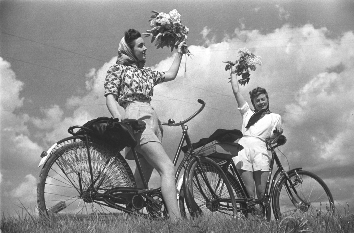 Damer på sykkeltur, her nyter de sol og lette skyer, samt nyplukkete blomster.