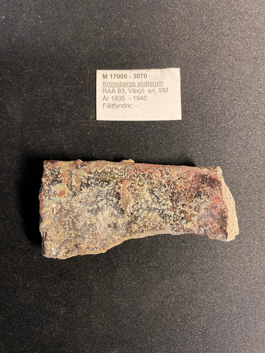 Fragment av en rak mynningsbit till en degel. Sekundärt bränd med delvis cintrade röda och gräna ytor på in- och utsidan. Relativt grovt gods. Liknar M 17000-3066.