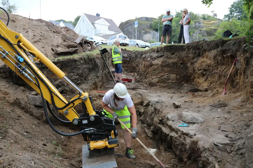 I et stort hull i bakken står en mann og graver med spade. Han har hvit hjelm på seg. Man ser deler av en gravemaskin i venstre del av bildet