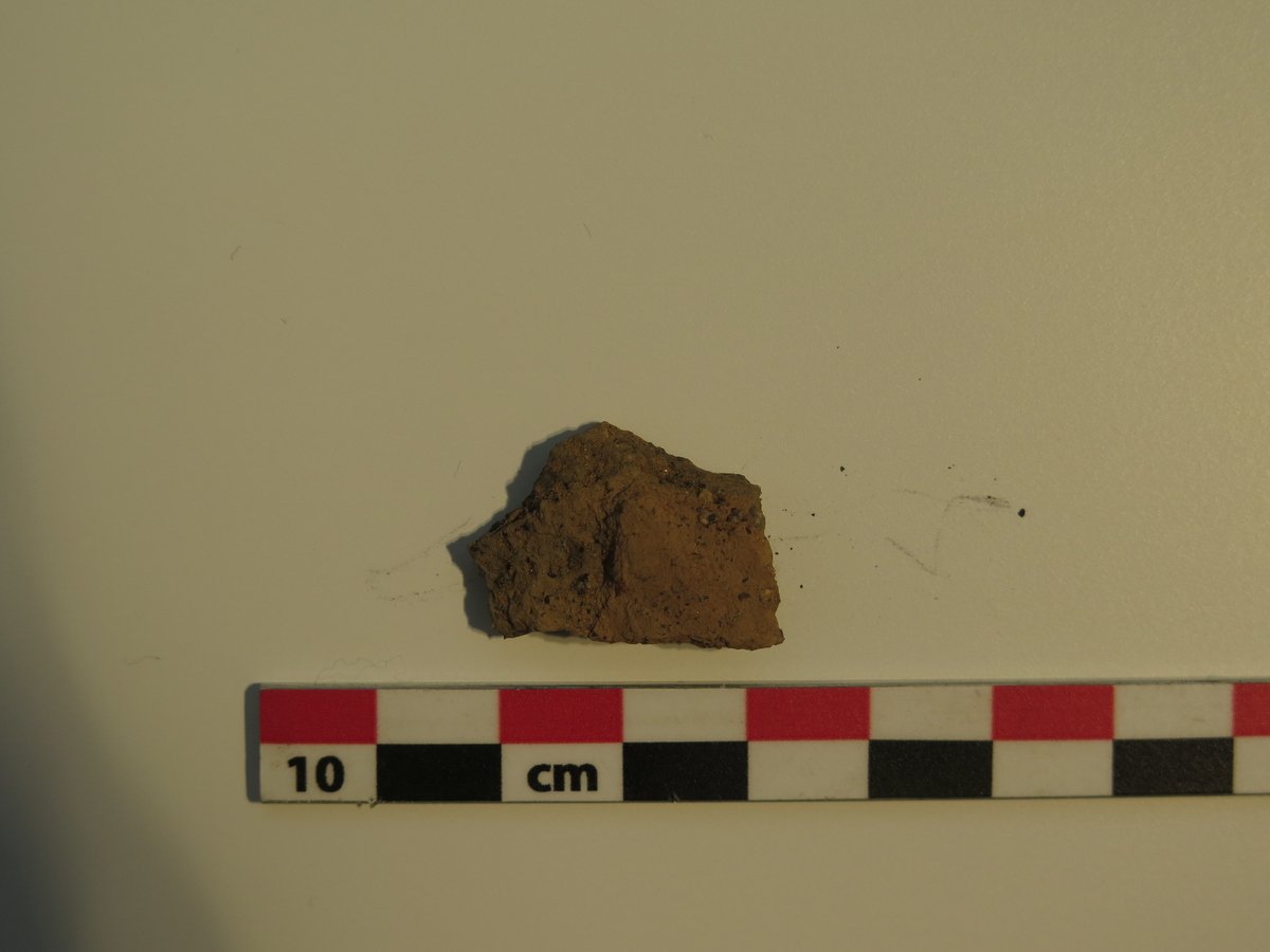 1 fragment fra kar av keramikk. Skåret var middels grovt magret, rødbrent på utsiden og mørk brunsort på innsiden. Vekt: 2,8 g. Stl.: 2,4 cm; Stb.: 1,8 cm; Stt.: 0,5 cm.
