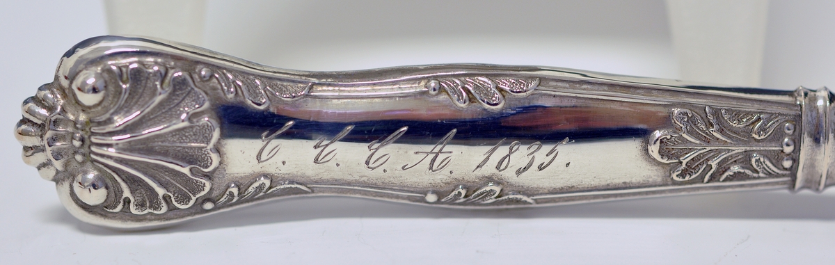 Kniv med skaft. Tilhører antakeligvis bestikksett med skje og gaffel. Bordkniv med butt knivblad. Skaftet har dekorert relieff i muslingmønster og qravert inn: C.C.C.A. 1835.(Christine Cecilie Charlotte Aall).