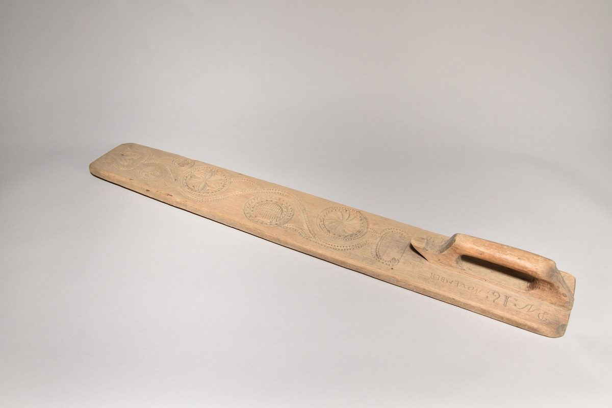 Kaveldon tillverkat i trä, med platt bräde samt skuret handtag. Brädets ovansida ornerat med karvsnitt samt med inskriften: "ANNO: 1723: SAD". 