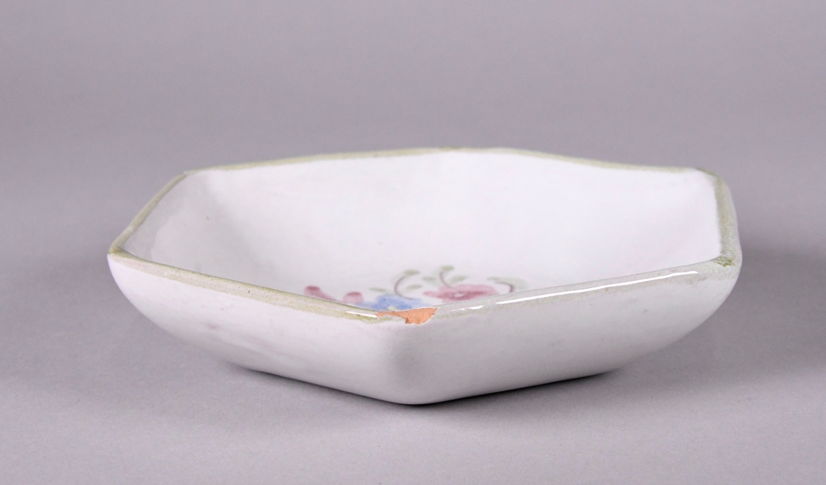 Skål av glassert keramikk, med opphøyd rand. Skålen er sekskantet, og har hvit grunnfarge. Håndmalt dekor av røde gule og blå blomster og en grønn borde langs randen.