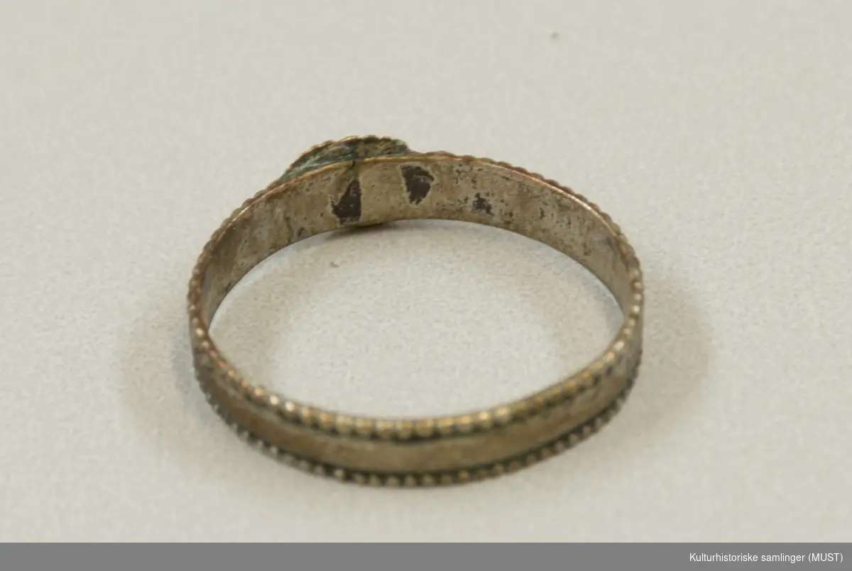 Glatt, flat ring med små, graverte border langs kantene. Påloddet ornament i
kobber bestående av to hender som holder hverandre.