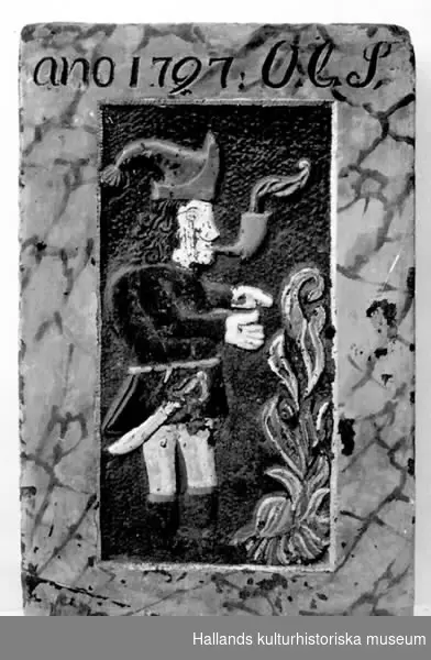 Sättugnssten av kalksten. Kortsidor och framsidans ram marmorerade i gulbrunt och svart. Rökande gubbe i relief, bemålad. Text: ANO 1797 O.C.S.
