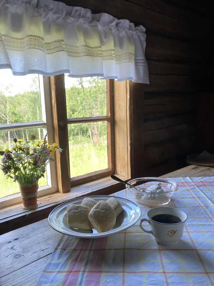 Bildet inne i Skålbergsætra. Brune tømmervegger. Sol skinner gjennom et vindu og lyser opp et bord. Villblomster i en vase i vinduskarmen. Godsaker og kaffe på bordet. Bordet har en rutete duk.