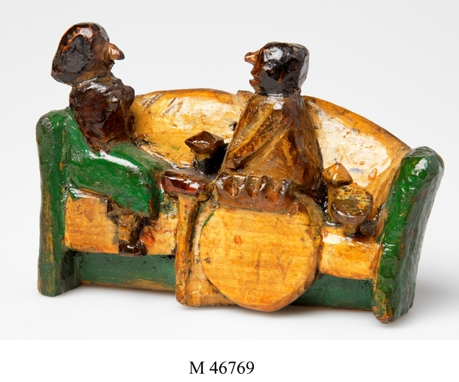 Trärelief. 
Utskuren trärelief. Målad i grönt och brunt.
Föreställer ett kafferep med två damer sittande i en
soffa med kaffebord bredvid sig.

Inskrivet i huvudbok 1985.
Montering/Ram: Ej ramad