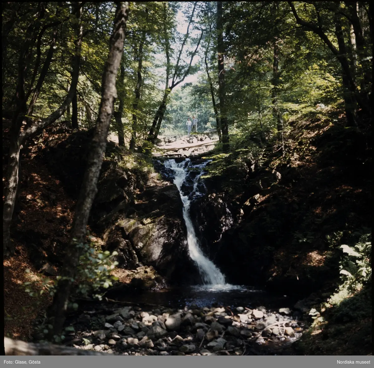 Ett vattenfall i ett vattendrag, en man och en kvinna står och tittar nedströms.
