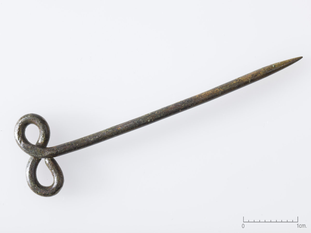 Nål av bronse, l.: 56 mm. Tenen bøyd til liggende S-formet hode, br.: 16 mm. Ten diam.: 2 mm. 
