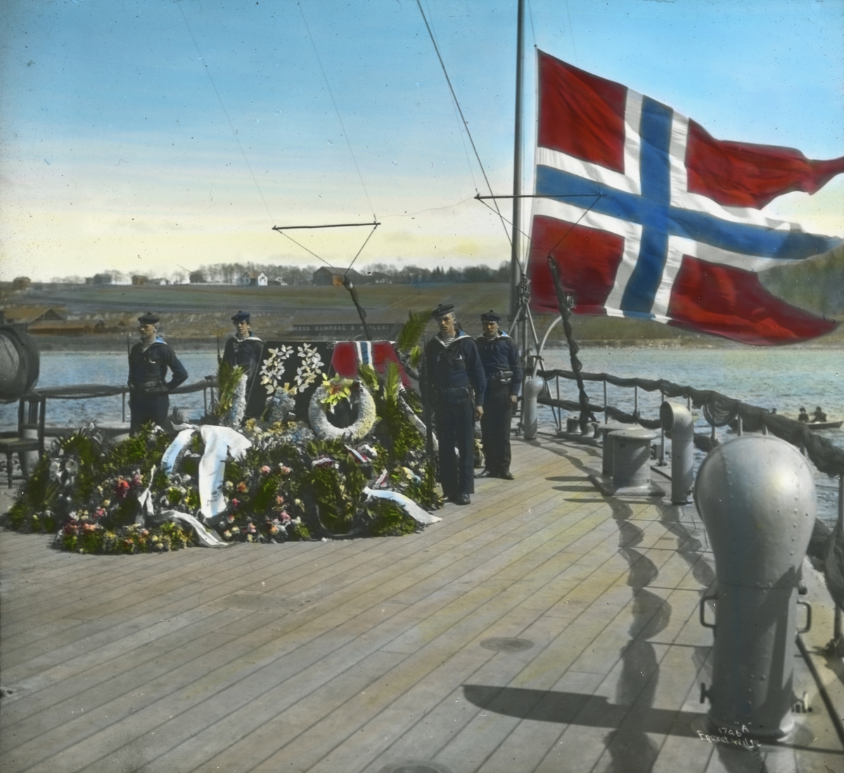 Håndkolorert dias. Bjørnstjerne Bjørnsons båre ombord i panserskipet Norge. 4 soldater står æresvakt og skipets flagg er firt på halv stang. Bjørnson døde i Paris den 26. april og ble begravet i Trefoldighetskirken den 3. mai 1910.