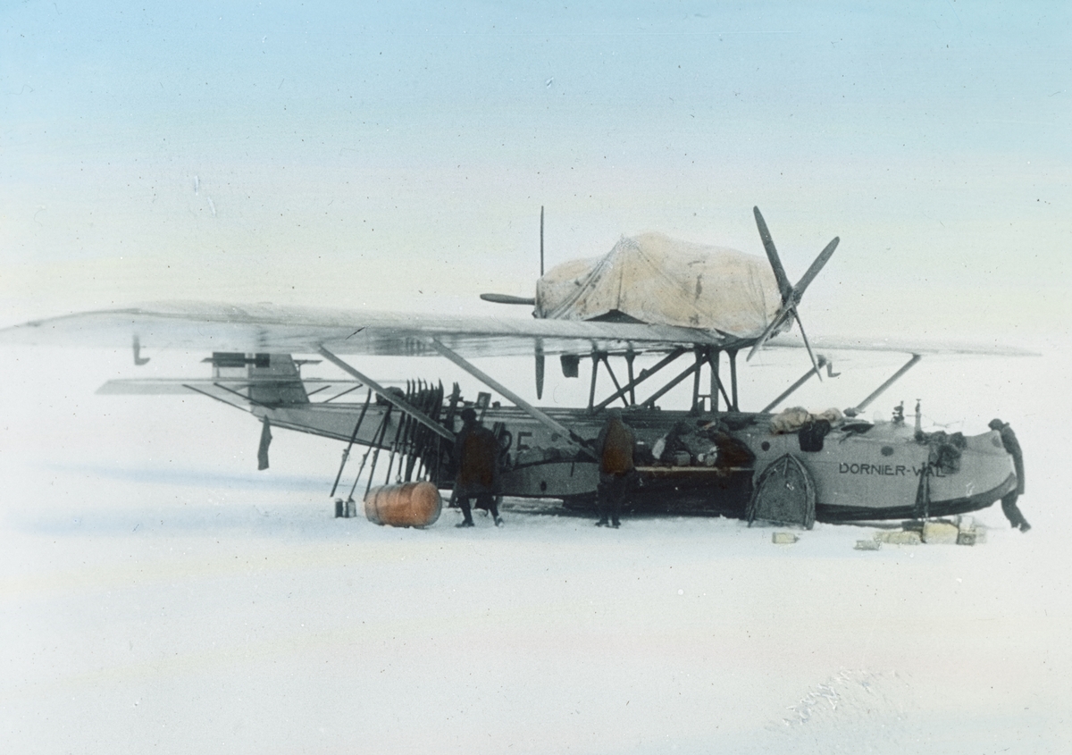 Håndkolorert dias. Dornier Do25 Wal N25 før starten fra Svalbard 21. mai 1925. Fire menn ordner med et fly. Lent inntil flykroppen står flere par ski.
