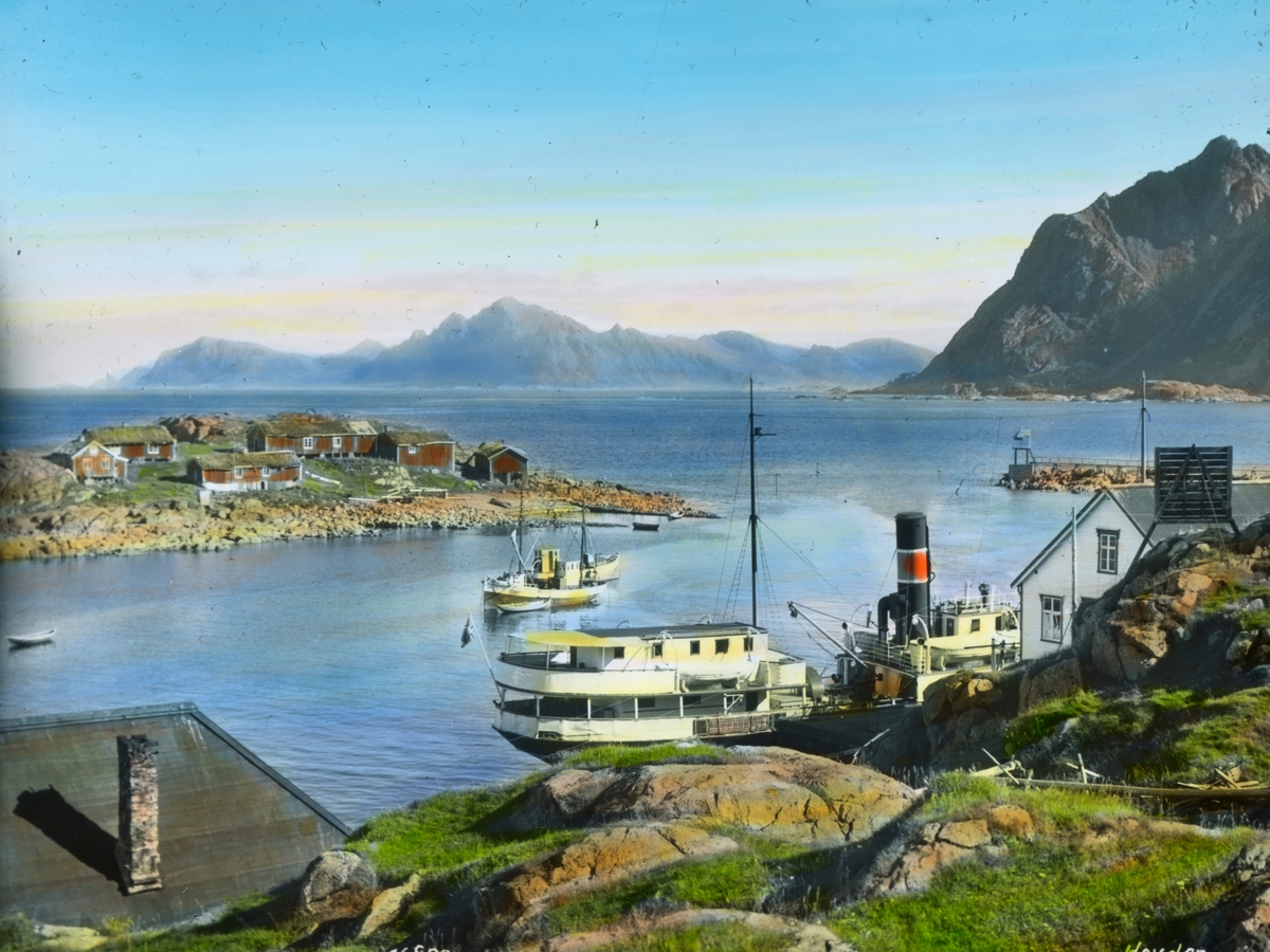 Håndkolorert dias. Fiskeværet Hovden i Vesterålen. Et dampskip ligger til kai. Bak sees to fiskebåter på fjorden og en øy kledd med små rødmalte hus.