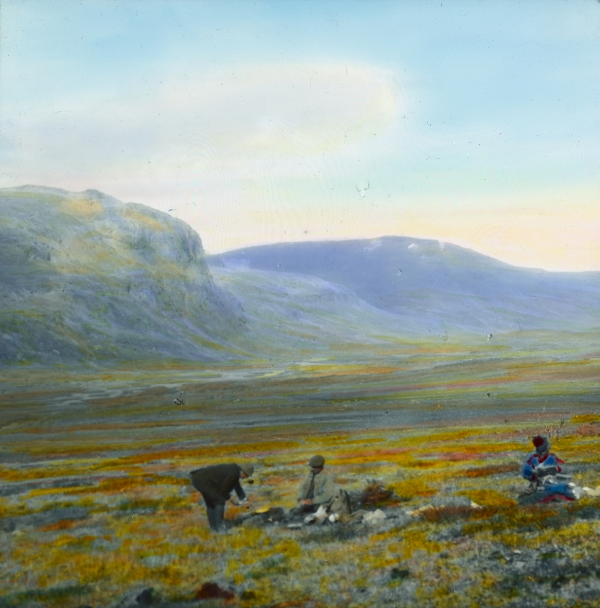 Håndkolorert dias. To menn, hvorav en antakelig er kledd i en samisk kofte, sitter på en høyfjellsslette eller vidde. En mann står ved et bål.