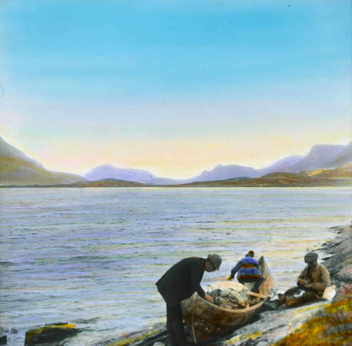 Håndkolorert dias. Tre menn ved en robåt i vannkanten. Den ene av mennene bærer samekofte og lue. I bakgrunnen ses fjell.