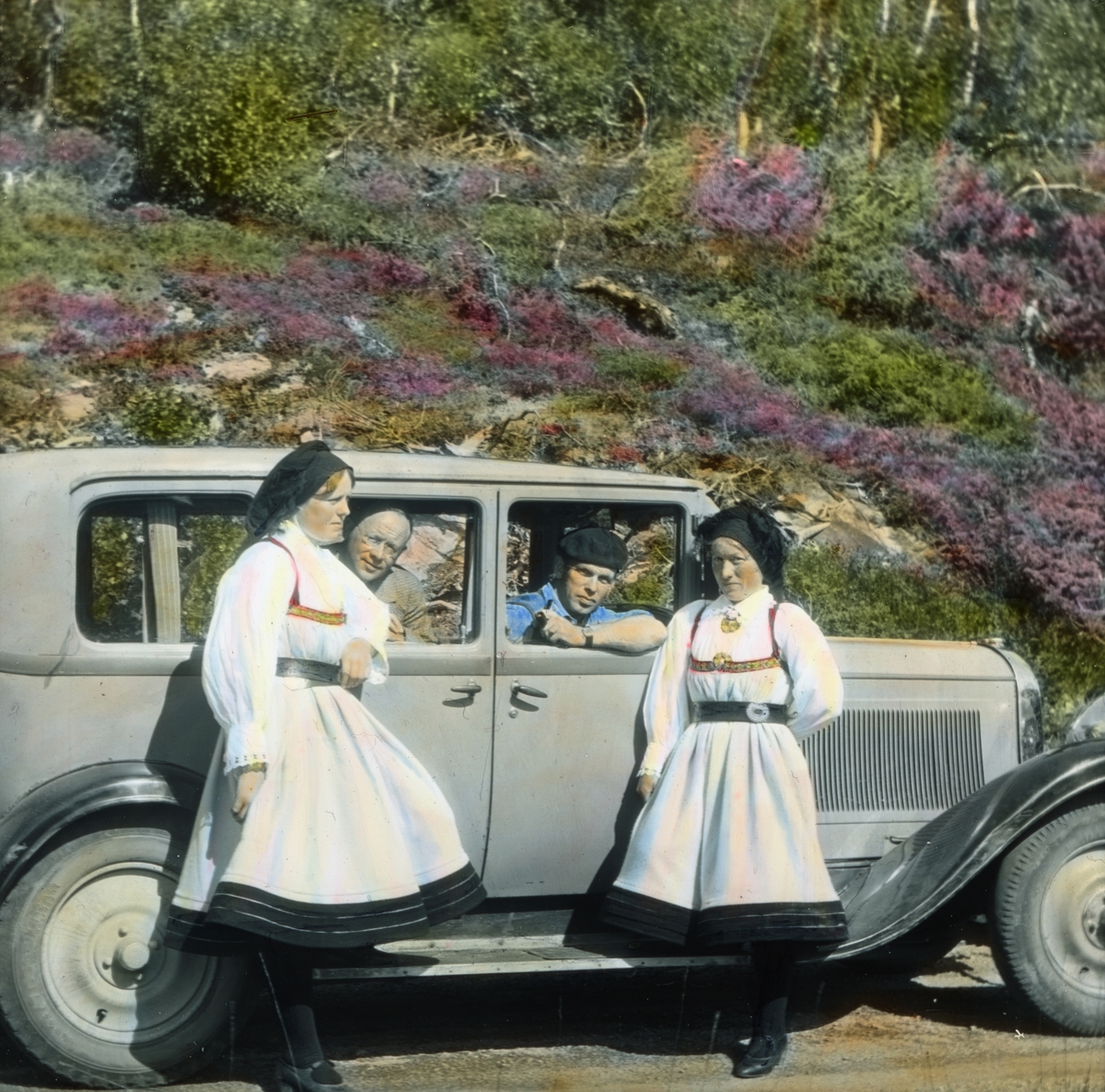 Håndkolorert dias. To kvinner i hvit setesdalsdrakt lener seg mot en bil, i bilen sitter to menn som titter ut av vinduene. Bilen er en 1929 Citröen SIX.
