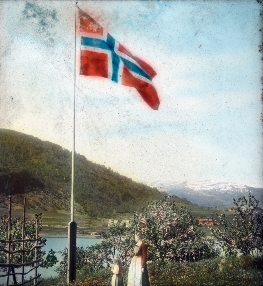 Håndkolorert dias. En kvinne og en jente i bunad står ved en flaggstang, der det norske flagget er heist.