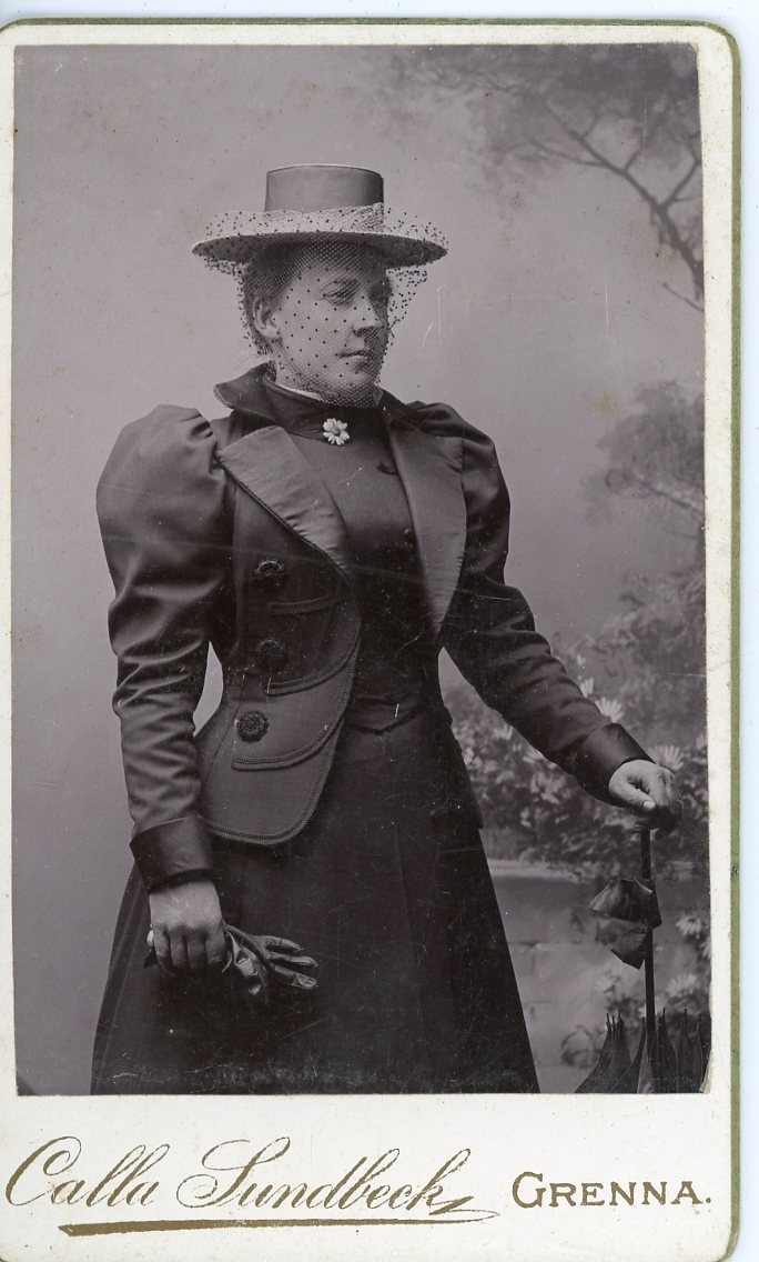 Kabinettsfotografi av en okänd kvinna som bär hatt med flor. I händerna har hon handskar respektive ett hopfällt parasoll.