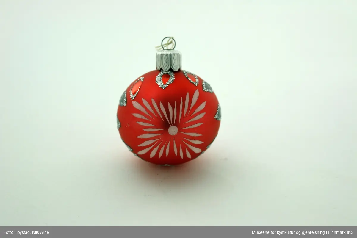 En liten julekule i hard plast med en halvmatt rød/mørkrosa farge med dekor. Dekoren består av påmalte hvite striper på begge sider av kulen og har et blomsterlignende mønster. I tillegg er det dråpeformet dekor malt rød og grønn med glitterkant som går vertikalt rundt kulen, og danner tilsammen en linje. Julekulen har et oppheng av metall med en hvit bomullstråd. Opphenget har gravert mønsterdekor.