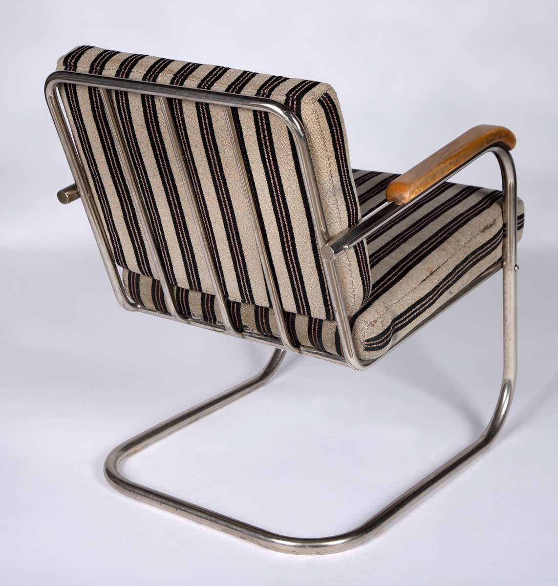 Stålrørslenestol med armlener i bjørk. Stolen er satt sammen av to stålrørselementer. Ben og lener er ett element og sete og rygg er ett element. Festepunkt midt på ryggen og foran på setet. Tre stålrørssprosser i ryggen og fjæring i sete. Gråbrunt møbeltrekk med vertikale brede, svarte striper som har to parallelle rader med smale vertikale striper. Disse er gråbrune med stiplete røde striper.