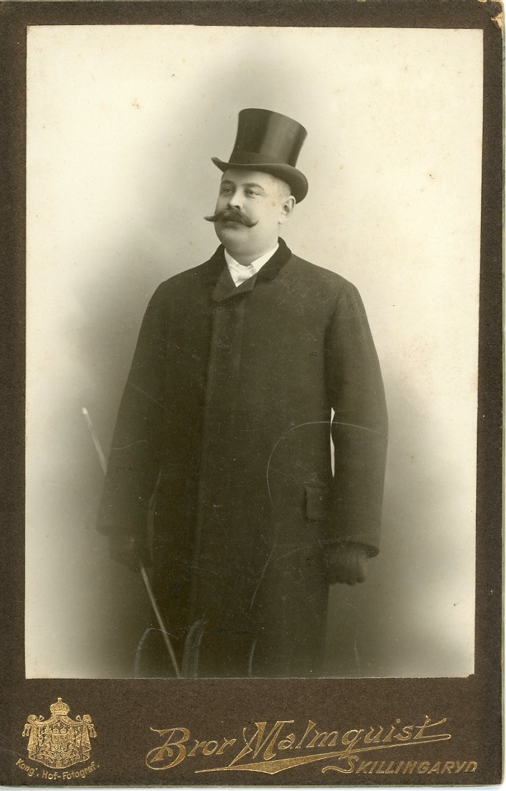 Kabinettsfotografi av en okänd man med mustasch. Han bär ytterkläder, hatt och en käpp i höger hand.